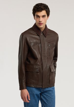 Junger Mann mit lockigem Haar, der eine braune Lederjacke mit Klappentaschen und einer Reißverschlusstasche an der Brust trägt, kombiniert mit blauen Jeans, stehend vor einem schlichten Hintergrund.