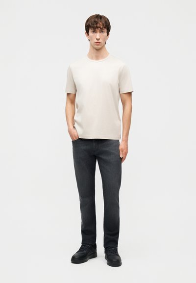 Νεαρός άνδρας όρθιος, φοράει απλό μπεζ κοντομάνικο T-shirt, σκούρα γκρι τζιν και μαύρα αθλητικά παπούτσια μπροστά σε μονόχρωμο λευκό φόντο.