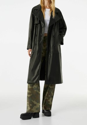 Woman wearing a long black leather trench coat over a white crop top, camo pants, and black platform shoes, standing against a plain background.
