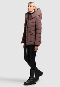 Brown padded jacket with a large hood, side zippers, and quilted design. Worn over a black top and paired with black leggings and boots.