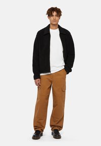 Schwarze Fleecejacke mit Kragen, darunter ein weißes Rundhals-T-Shirt, braune Cargo-Hosen mit Seitentaschen und schwarze Schnürschuhe.