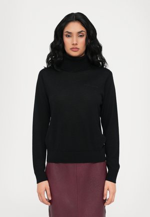 Svart genser med høy hals laget av myk stoff med lange ermer. Har en subtil merkevarelogo og en tettsittende design. Kombinert med et burgunder lærskjørt.