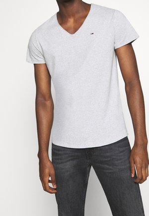 Mężczyzna ubrany w jasnoszarą koszulkę z dekoltem w serek, z małym logo Tommy Hilfiger na piersi oraz ciemnoszare dżinsy, stojący na białym tle.