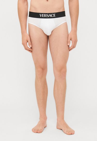 Bílé pánské slipy s černým elastickým pasem zdobeným logem "Versace". Umístěny na nahém modelu na jednobarevném pozadí.