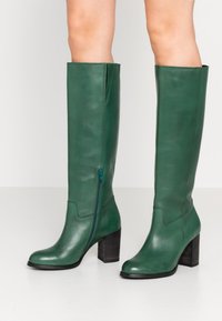 Bottes montantes en cuir vert avec une fermeture éclair latérale et un talon carré. Texture lisse, bout arrondi, et détails cousus en haut.