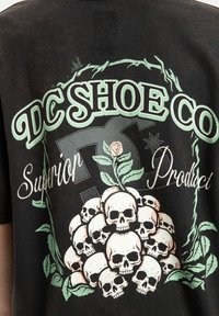 Čierne tričko má grafický dizajn s vrstvami lebiek obklopených zelenými viničmi. Text obsahuje "DC Shoe Co." a "Superior Product" s opotrebovaným vzhľadom.