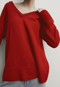 Pull en maille rouge avec un décolleté en V profond, des poignets et un ourlet côtelés, présentant une coupe ample et une texture douce. Idéal pour un look décontracté.