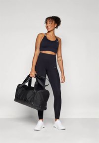 Sort sports-bh, sorte leggings med højtaljet pasform og hvide sportssko. Personen holder en sort duffel taske med to håndtag.