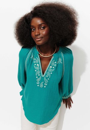 Femme aux cheveux bouclés volumineux portant un chemisier teal brodé et un collier en or, debout devant un fond blanc.