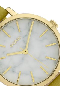 Gold-tone watch featuring a marbled white dial, gold hour markers, and a light green leather strap. Minimalist design with clear time indicators.