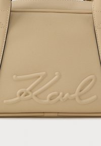 Bežs ādas somas panelis ar izšūtām rokturiem un uzrakstu "Karl" reljefā centrā.