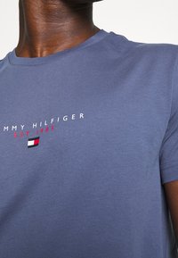 Námořnická modrá bavlněná tričko s kulatým výstřihem. Obsahuje bílý text "TOMMY HILFIGER" a rok založení 1985, doplněný malým logem.