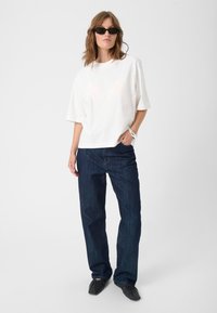 White oversized t-shirt with short sleeves, paired with dark blue straight-leg jeans. Model wearing black sunglasses and flat black shoes.