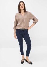 Beige Satinbluse mit V-Ausschnitt und gerafften Ärmeln, kombiniert mit navy-blauen, eng anliegenden Hosen. Akzentuiert mit einem schwarzen Gürtel und schwarzen Netzschuhen.