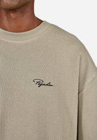 Zbliżenie osoby w beżowym swetrze z prążkowanej dzianiny z czarnym, kursywą napisem "Pegador" na klatce piersiowej, na jednolitym jasnym tle.