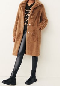 Manteau en fausse fourrure marron avec un large col, des boutons à l'avant et des poches latérales, associé à un legging en fausse cuir noir et des bottes à la cheville.