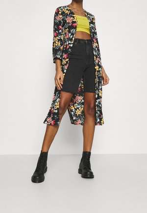 Personne portant un long manteau noir à motifs floraux, un top court jaune, un short noir taille haute et des bottines noires à lacets, debout sur un sol gris.