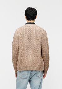 Άτομο που φαίνεται από πίσω, φοράει μπεζ πουλόβερ πλεκτό με σχέδιο καλώδιο πάνω από μαύρο πουκάμισο, συνδυασμένο με ανοιχτό μπλε τζιν τζινς, σε απλό φ όντο.