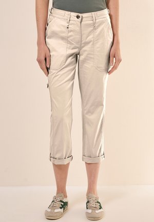 Beige Hosen mit Bündchen, vorne und seitlich Taschen, getragen mit grauen und grünen Sneakers.