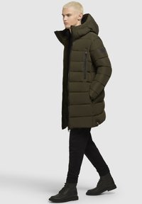 Olivno zeleni pufer jakna z velikim kapuco, horizontalno šivana, zadrgo stranskega žepa in črnimi detajli, nosi z črnimi hlačami in čevlji.