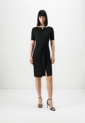 Woman standing in a black knee-length dress with short sleeves, a front slit, belt tied at the waist, and black strappy high heels.