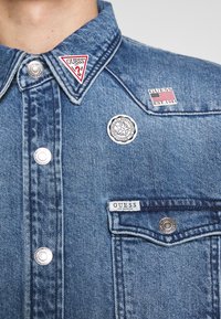 Vaate takki vaaleansinisestä farkusta, jossa on metallinapit ja erilaisia tägejä: kolmion muotoinen punainen Guess-logo, musta pyöreä emblem ja Guess-eteinen.