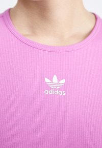 Růžové tričko s žebrovaným vzorem a kulatým výstřihem, na přední straně s bílým logem Adidas a třemi pruhy.