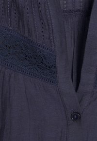 Blusa blu marina con tessuto testurizzato, cuciture verticali e un pannello di pizzo decorativo. Presenta una chiusura con bottoni frontale e un morbido drappeggio.