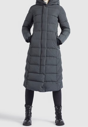 Mujer con un abrigo largo acolchado de invierno en gris oscuro, con capucha, cremallera frontal y bolsillos laterales, combinado con leggings negros y botas negras con cordones.