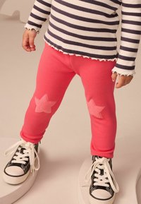 Barn i randig långärmad tröja och rosa leggings med stjärnlappar, som står i svarta sneakers på en enkel vit pedestalbakgrund.