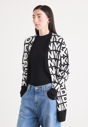 Cardigan bianco e nero con lettere geometriche audaci, materiale lavorato a maglia, apertura frontale, maniche lunghe, abbinato a un top nero e jeans blu.