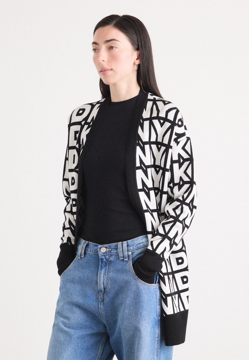 Cardigan preto e branco com letras geométricas em negrito, material tricotado, frente aberta, mangas longas, combinado com uma blusa preta e jeans azuis.