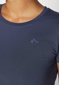 Camisa de manga corta de color azul marino, confeccionada en una tela lisa y elástica; presenta un escote redondo y un logo sutil con acentos circulares en el pecho.
