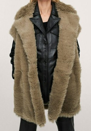 Model wearing sleeveless tan faux fur vest over black buttoned leather jacket and black pants against plain background.