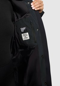 Chaqueta negra con un interior de satén suave, cierres a presión y etiquetas de tela que indican "Khujo" e instrucciones de cuidado.