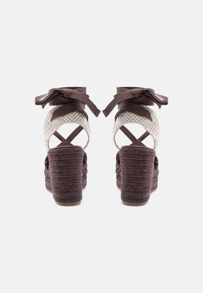 Paire de sandales compensées marron avec talons tissés, brides de cheville blanches tissées et rubans marron noués autour de la cheville, vues de dos.