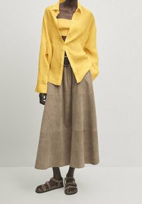 Chemise en lin jaune boutonnée en haut, associée à une longue jupe en daim brun et des sandales à brides marron. Design simple avec une coupe décontractée.