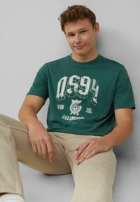 Groene katoenen t-shirt met een groot wit grafisch ontwerp met "QS94" en een wereldbolmotief. Model zittend, gekleed in beige broek. Gladde textuur.