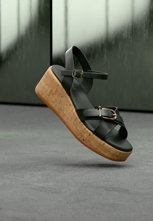 Svart skinnplattform sandal med korksåle, med to gullspenner og ankelstropp, svevende over et grått gulv.