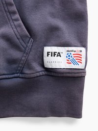 Σκούρο γκρι ύφασμα με λευκή ετικέτα FIFA Classics που διαθέτει μπλε ποδοσφαιρική μπάλα και κόκκινες λωρίδες με κείμενο "World Cup USA 94."