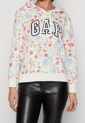 Kobieta ubrana w biały, kwiatowy kaptur z dużym napisem „GAP” oraz czarne skórzane spodnie, widoczna od ramion do bioder.