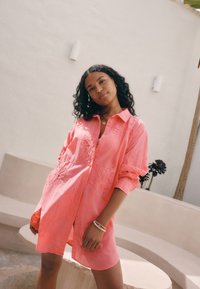 Rožinės spalvos siuvinėta marškinėliai su gėlių raštais, laisvo kirpimo, ilgomis rankovėmis. Švelnios tekstūros audinys, turi apykaklę ir šonines skeltukes.