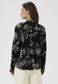 Frau mit langen braunen Haaren, die eine schwarze blumige Bluse mit langen Ärmeln und hellbeigen Hosen trägt. Sie steht mit dem Rücken zu einem schlichten hellen Hintergrund.