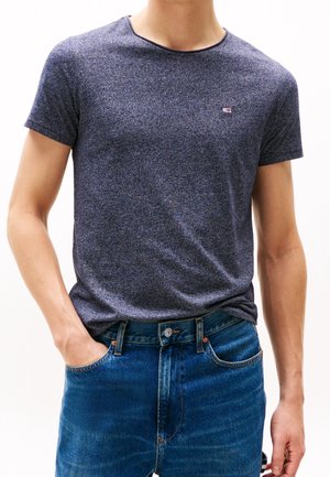 Marineblaue T-Shirt aus strukturiertem Stoff mit kurzen Ärmeln und rundem Ausschnitt, getragen mit blauen Denim-Jeans, Hand in der Tasche. Kleines Logo sichtbar.