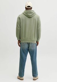 Grön hoodie med huva baktill, avslappnad passform och ribbade muddar, kombinerad med ljusblå denimjeans och beige skor. Enfärgad bakgrund.