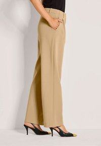 Vrouw in beige wijde broek en zwarte slingback hakken met gouden teentips, staand met één hand in haar zak tegen een effen achtergrond.