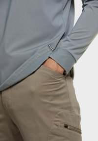 Lichtgrijze lange mouw top met zijsplit en contrasterende zoom, gecombineerd met beige broeken met een ritszak en een gladde textuur.