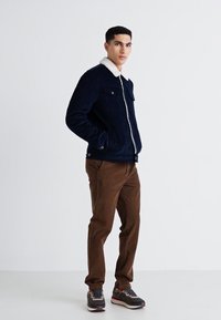 Granatowa kurtka z korduroju z białym kołnierzem z polaru, zapinana na zamek, z dwiema kieszeniami na piersi. Połączona z brązowymi spodniami i wielokolorowymi trampkami.