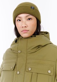Vrouw in een olijfgroen beanie en een bijpassende capuchonjas met grote knoopzakken, kijkend naar de camera tegen een witte achtergrond.