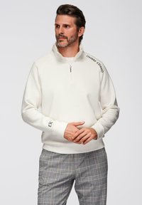 Sudadera blanca con cuello de media cremallera, con texto negro en la manga. Combinada con pantalones de cuadros grises, que muestran una textura suave.